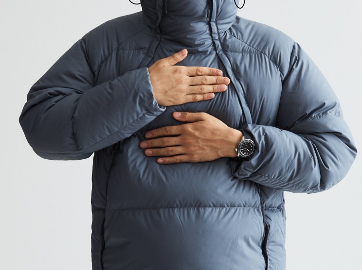 Man wearing gray puffy down jacket with hood up, hands crossed over chest, wearing black watch