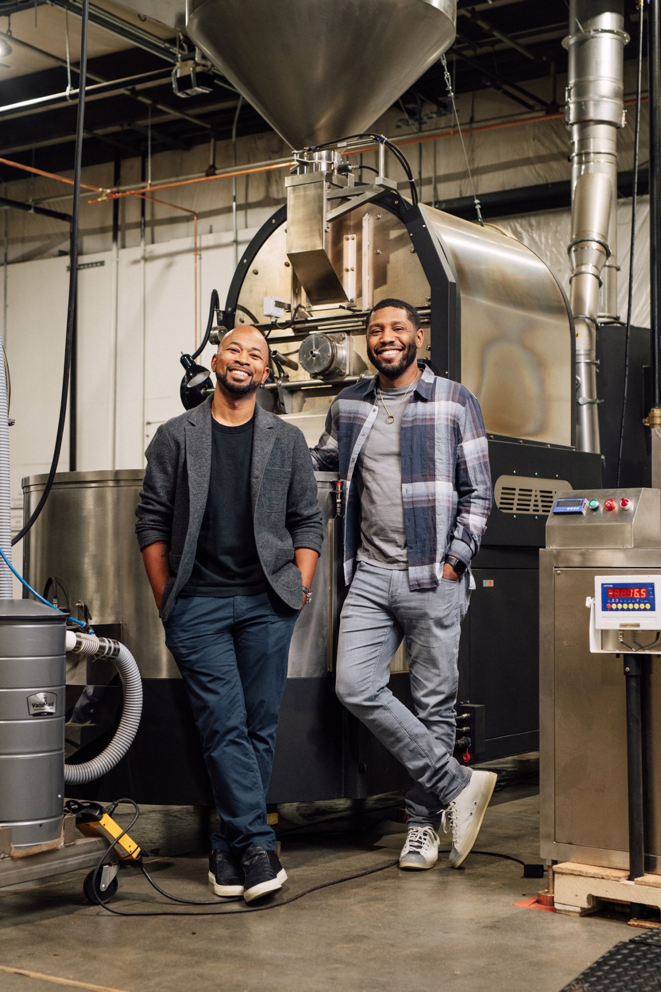 Pernell Cezar and Rod Johnson standing in front of a large commercial coffee roasting machine in their Blk & Bold roastery facility.