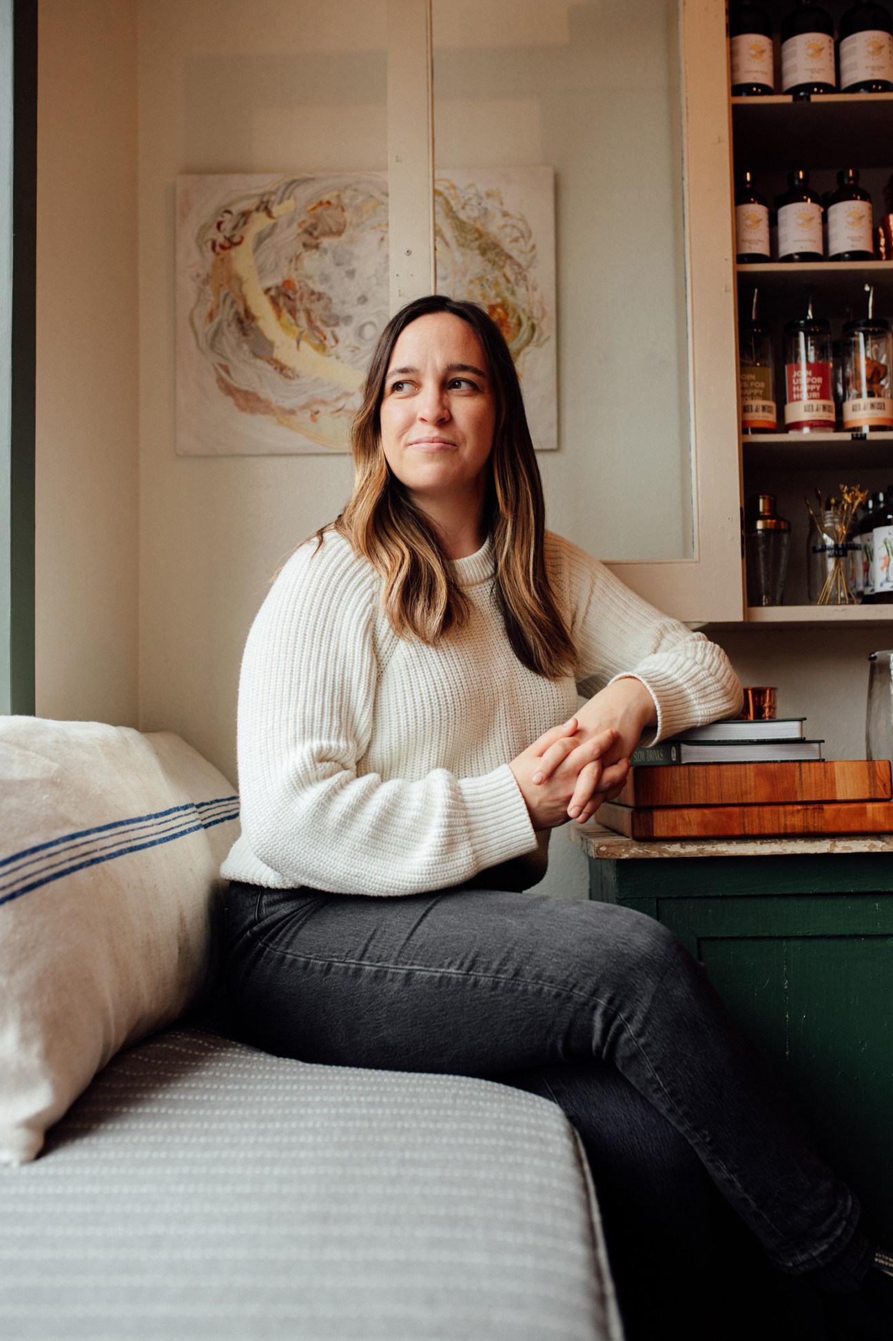 Woman sitting on a sofa next to stacked books and shelving with bottles, looking away from camera in neutral setting.