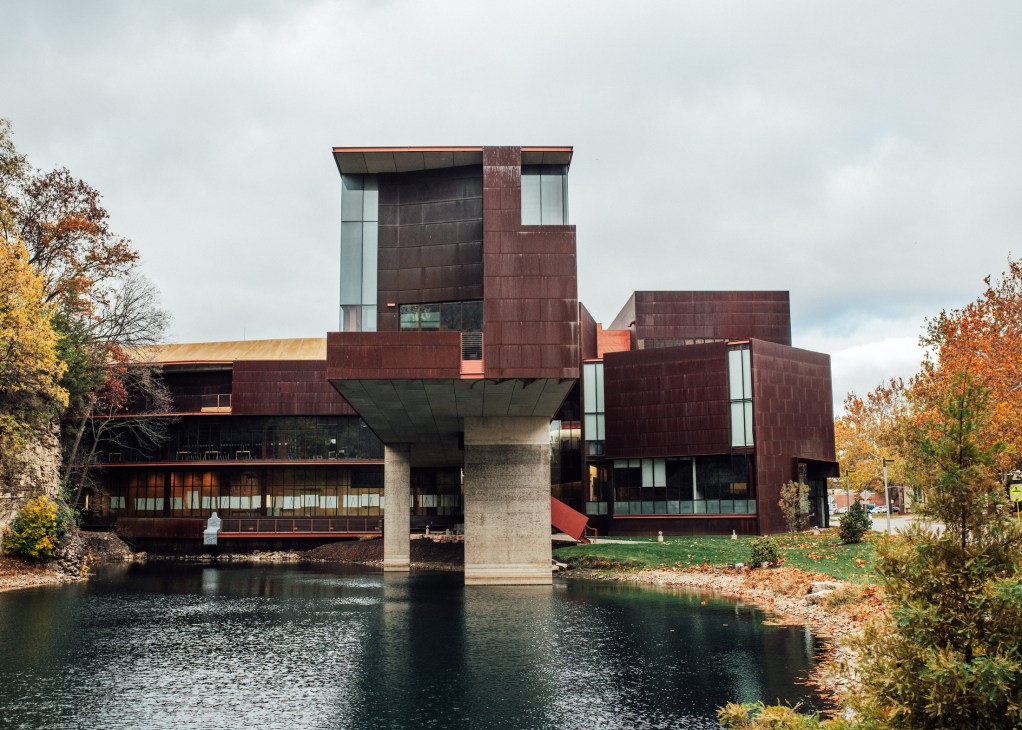Modern art building with burgundy metal panels beside river in autumn setting.