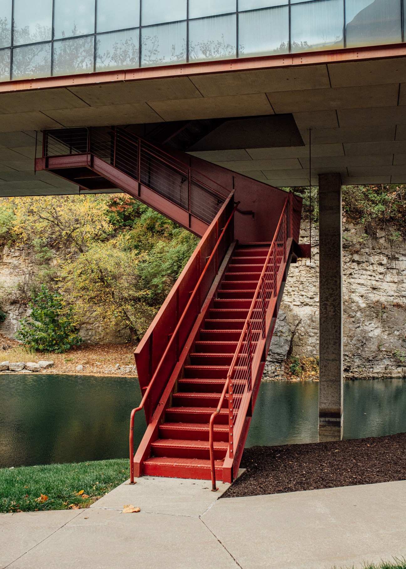 Red metal staircase descending from a modernist concrete building to a riverbank with glass windows above.