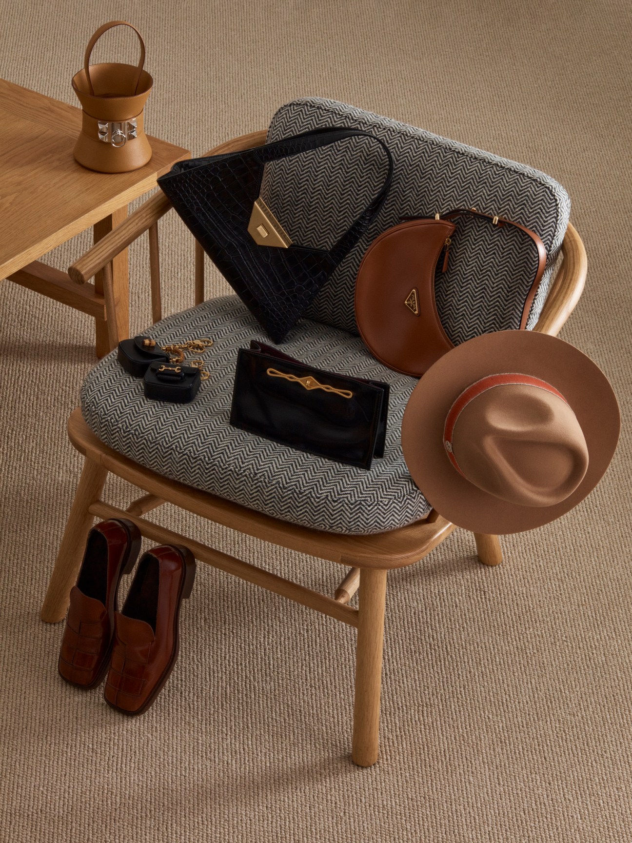 Luxury accessories arranged on a wooden chair with herringbone blanket, including Hermès, Bally and Prada bags, Maison Michel hat, and Gucci charms.