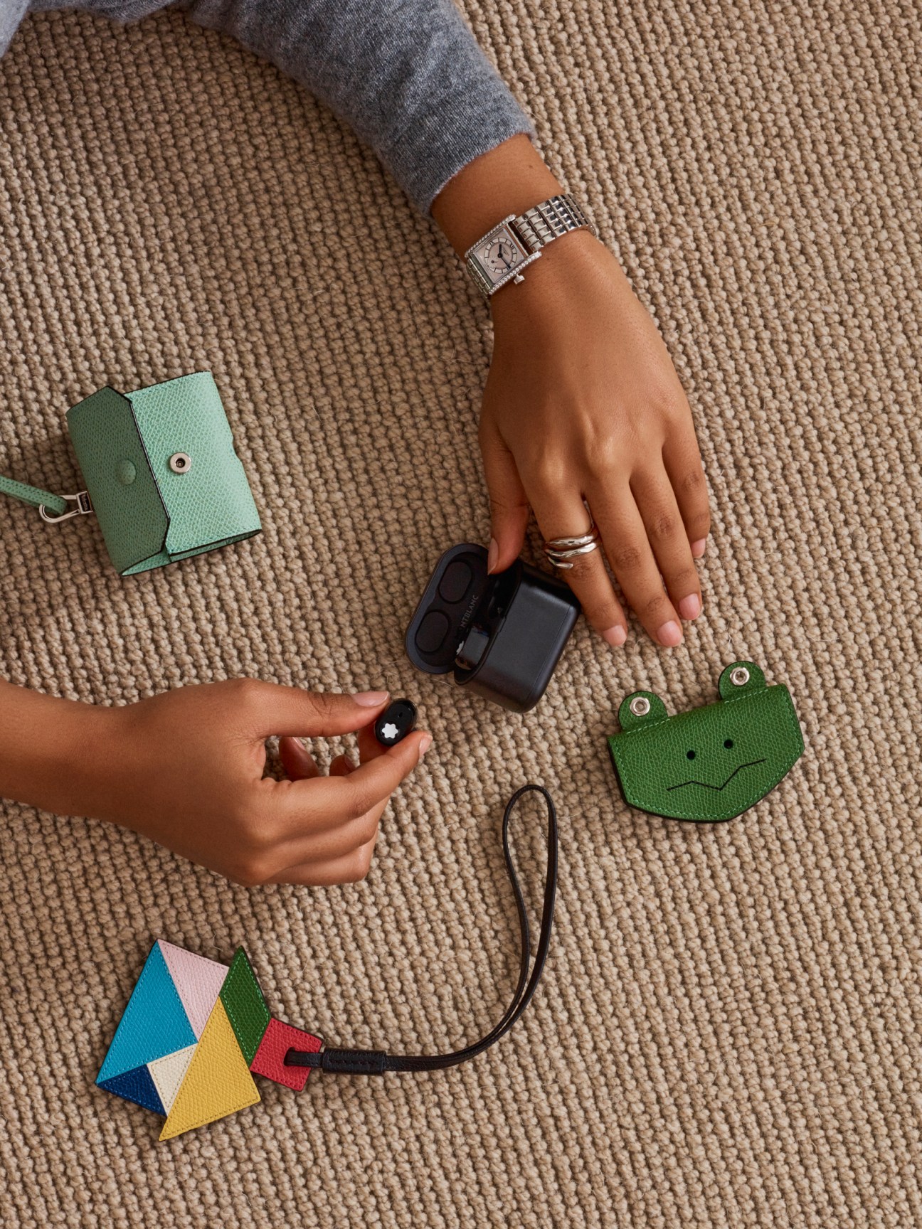 Woman's hands displaying Longines watch, Goossens ring, Valextra accessories and Montblanc headphones on textured fabric.