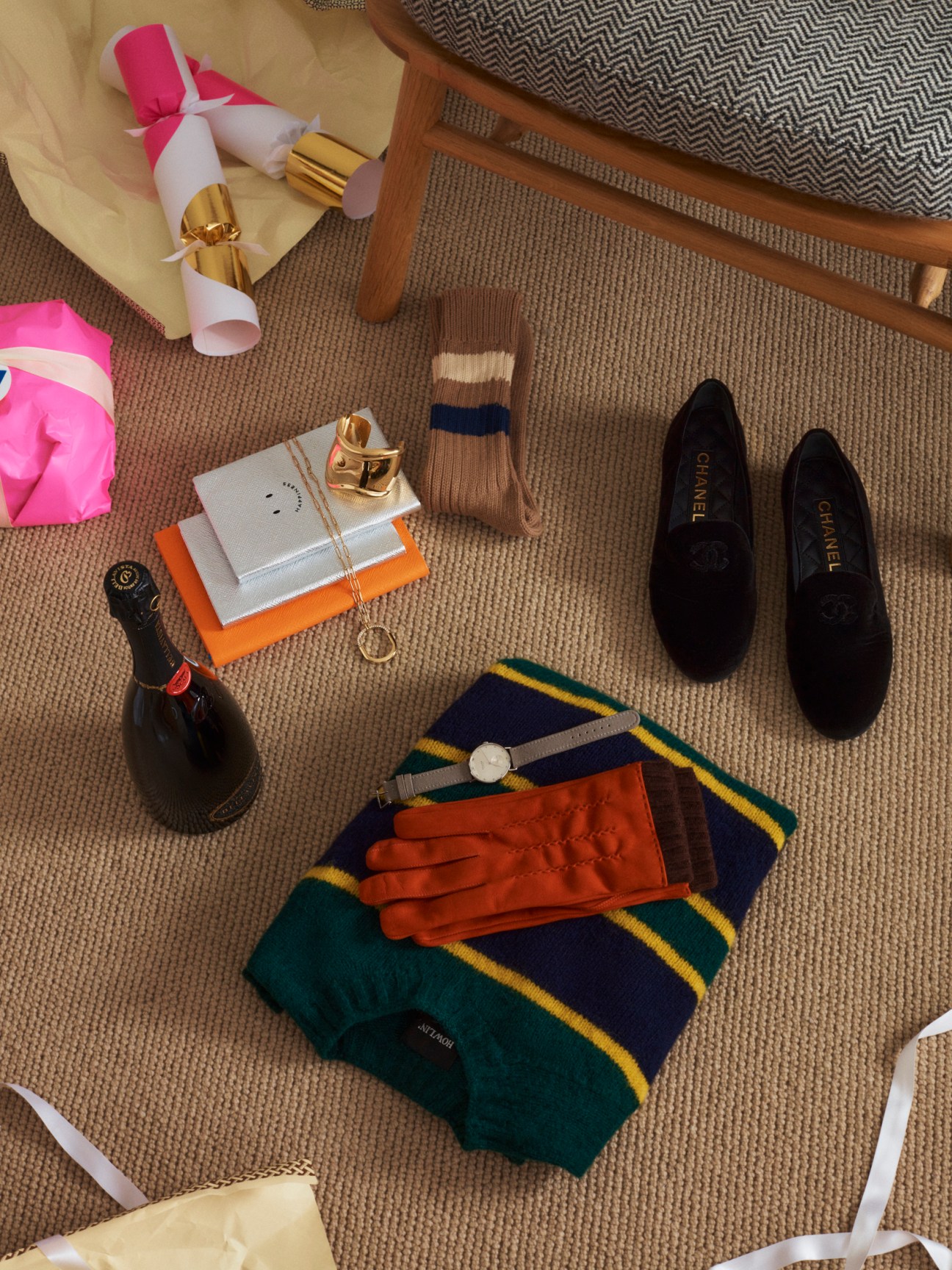 Luxury gift items arranged on carpet including Chanel shoes, Thomas Riemer gloves, Nomos watch, champagne bottle and wrapped gifts.