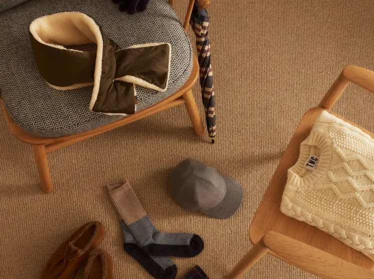 Overhead view of winter accessories including knit sweater, tartan scarf, wool socks, and suede shoes on neutral carpet