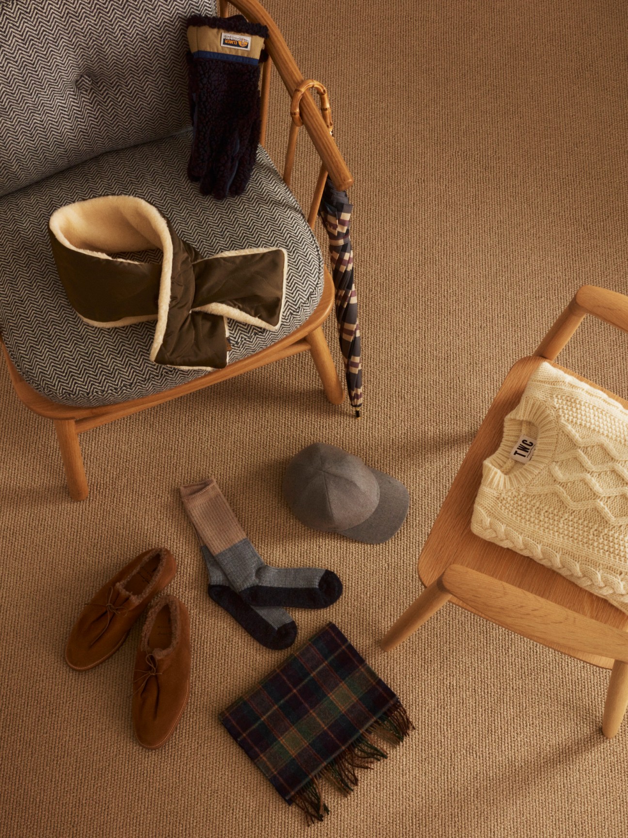 Flat lay of classic menswear items including a jumper, scarves, socks, shoes, cap, gloves, and umbrella arranged on a textured beige background.