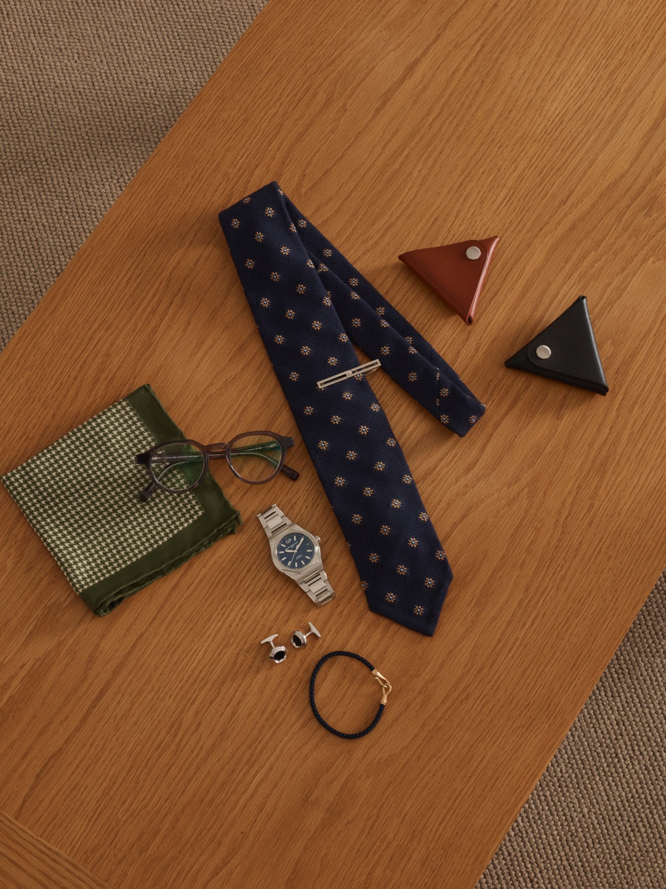 Flatlay of menswear accessories including navy patterned tie, glasses, leather bag charms, metal watch, cufflinks and bracelet on wooden surface.