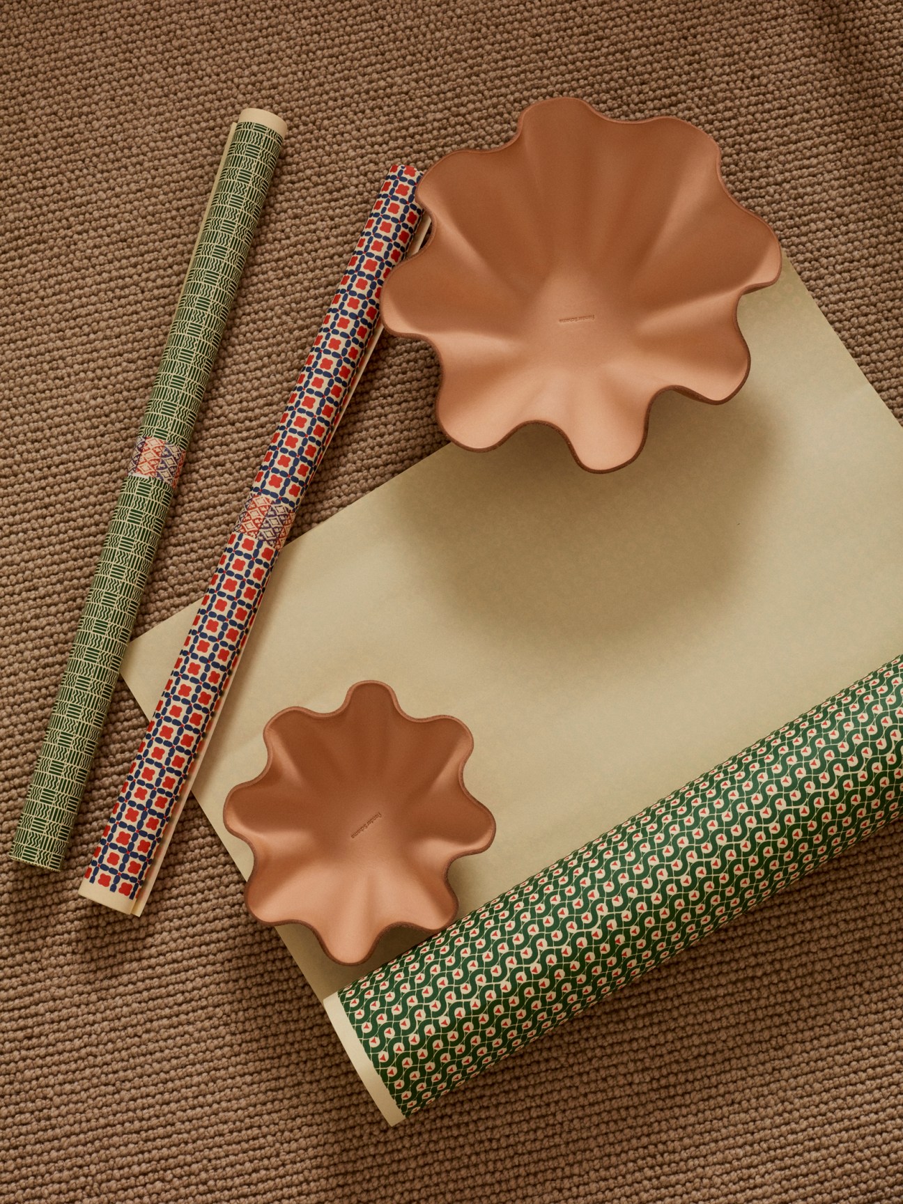 Two terracotta shell-shaped bowls displayed on patterned wrapping papers in neutral and green tones.