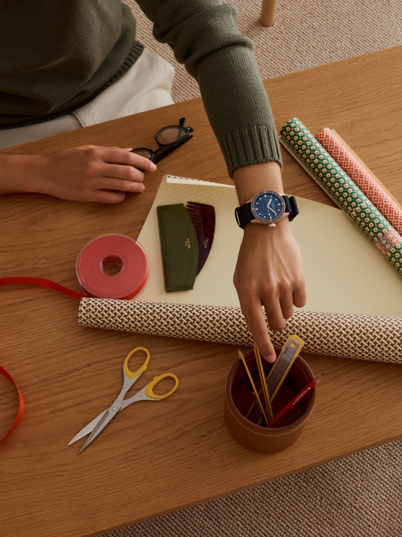 Person wearing a green jumper and watch arranging gift-wrapping supplies on a wooden desk.