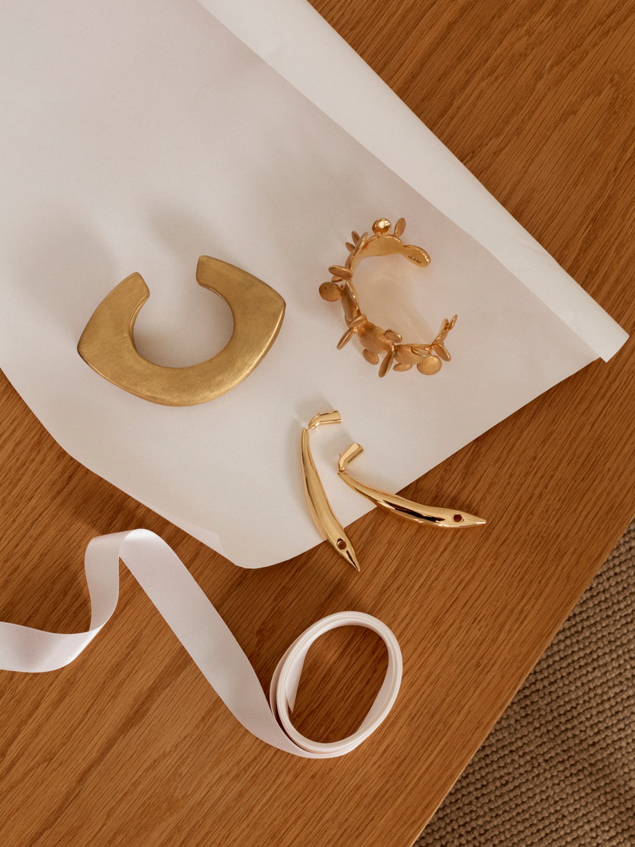 Gold cuffs, Bottega Veneta earrings and white ribbon displayed on white paper against wooden surface.