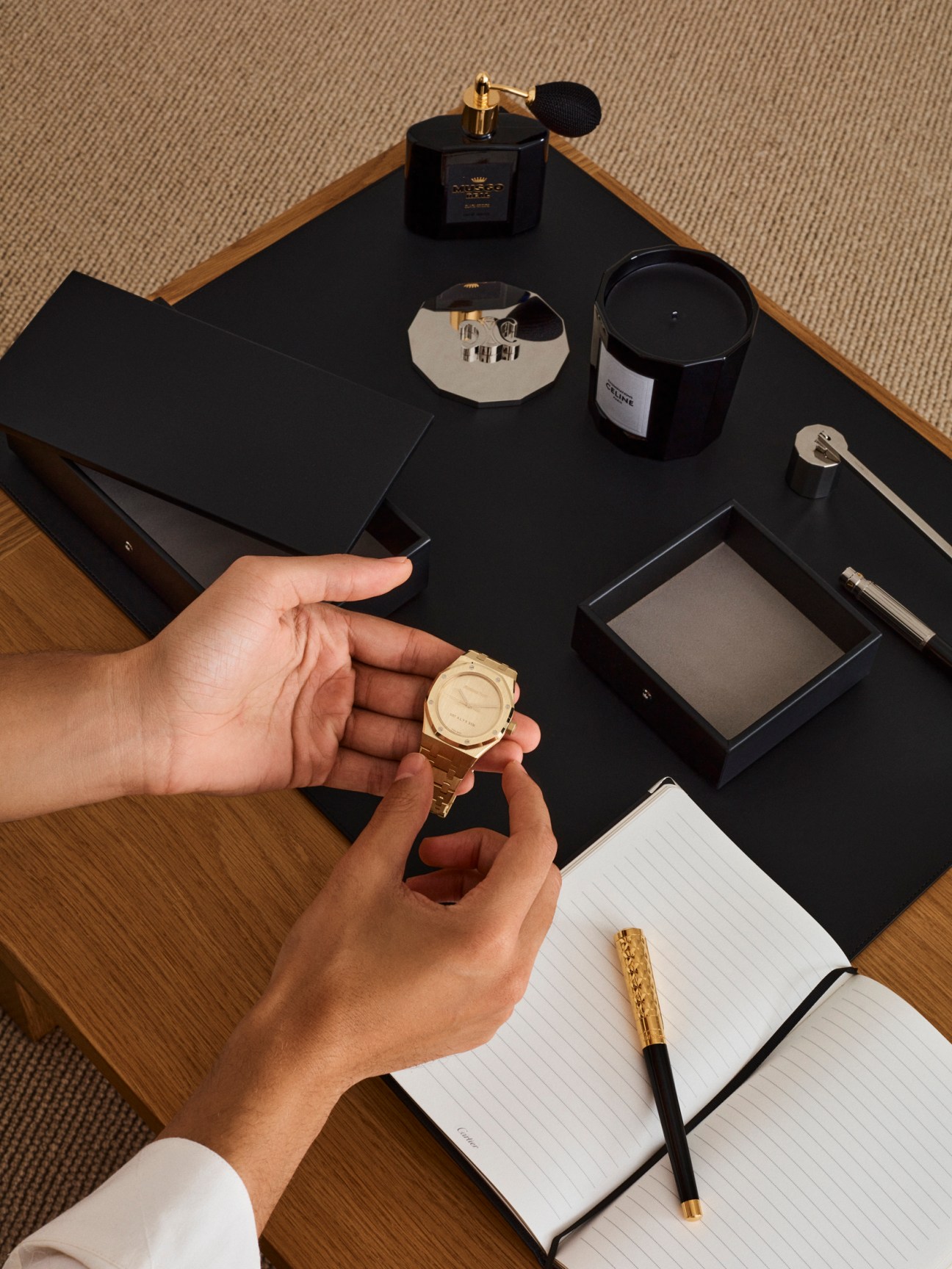 Hands holding a gold watch displayed on a desk with luxury desk accessories including pen, notebook, candle, and fragrance.