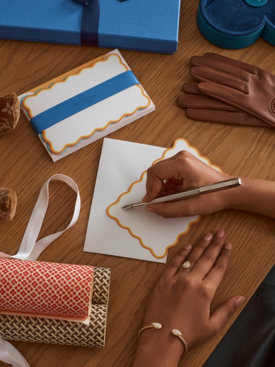 Hands writing on decorative stationery with scalloped edges, surrounded by leather gloves and gift boxes