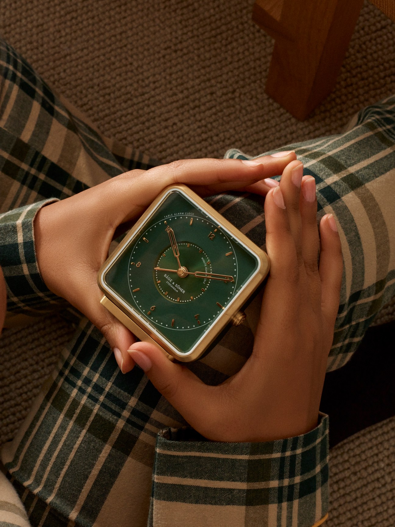 Hands holding a square green table alarm clock with gold trim and copper accents over a plaid blanket.