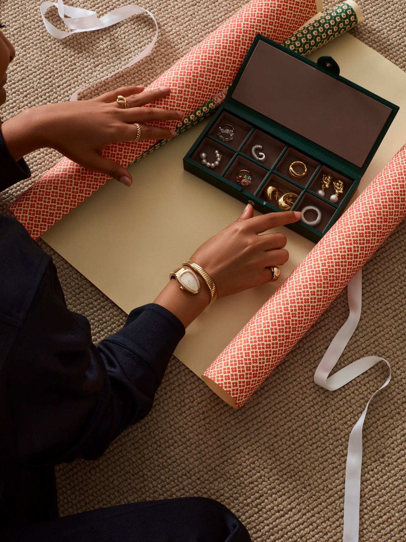 Hands displaying jewelry in an open green box with red patterned wrapping paper and ribbon.