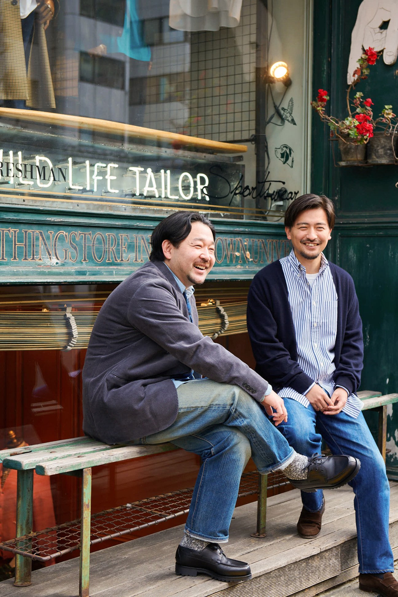 Two men sitting outside Wild Life Tailor shop in Tokyo, smiling at the camera.