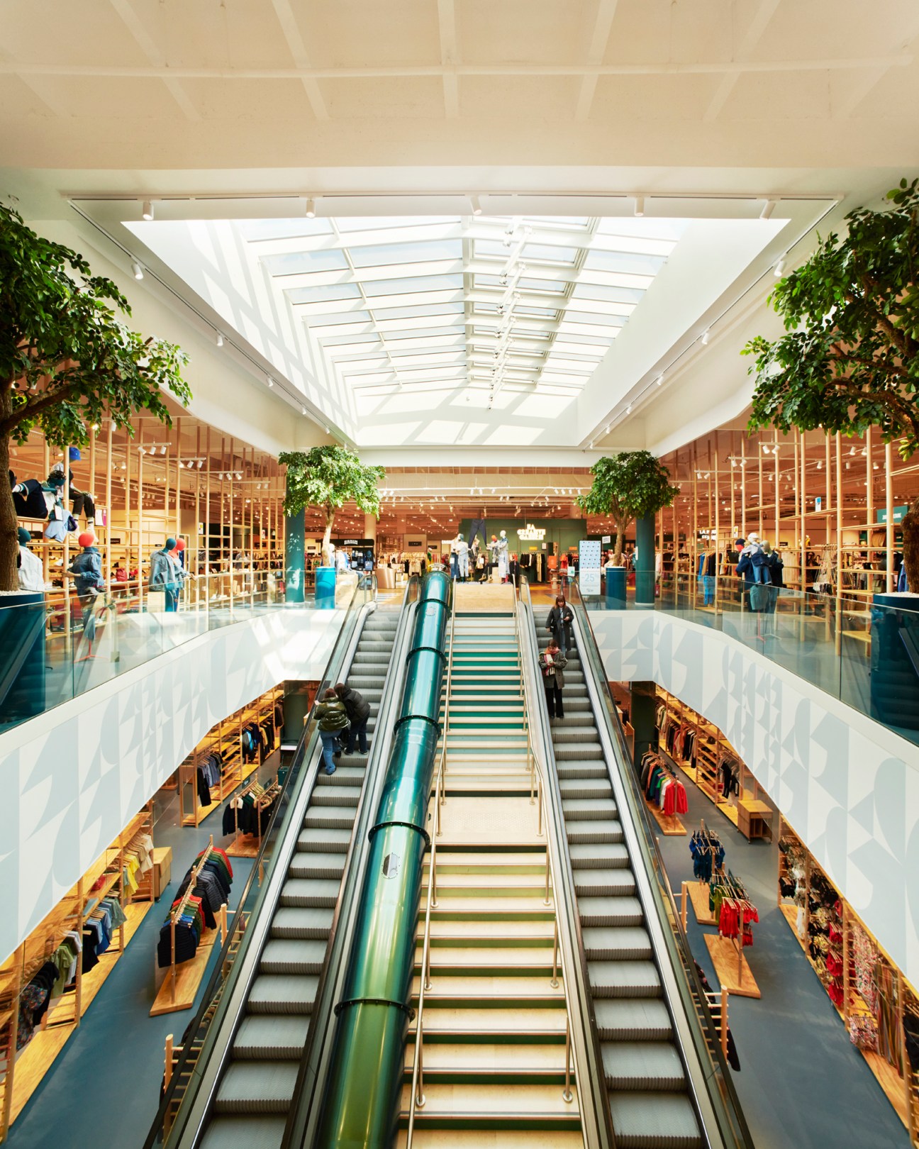 Modern shopping center interior with escalators, skylight, and multi-level retail spaces with wooden fixtures