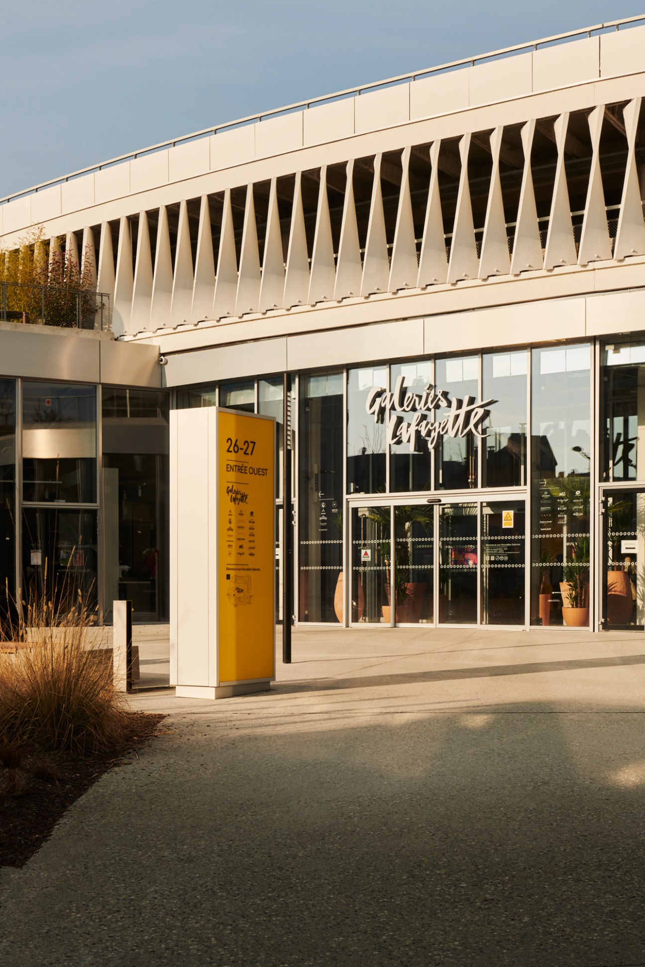 Yellow wayfinding sign reading '26-27 ENTREE OUEST' outside modern building with glass facade and geometric white overhang