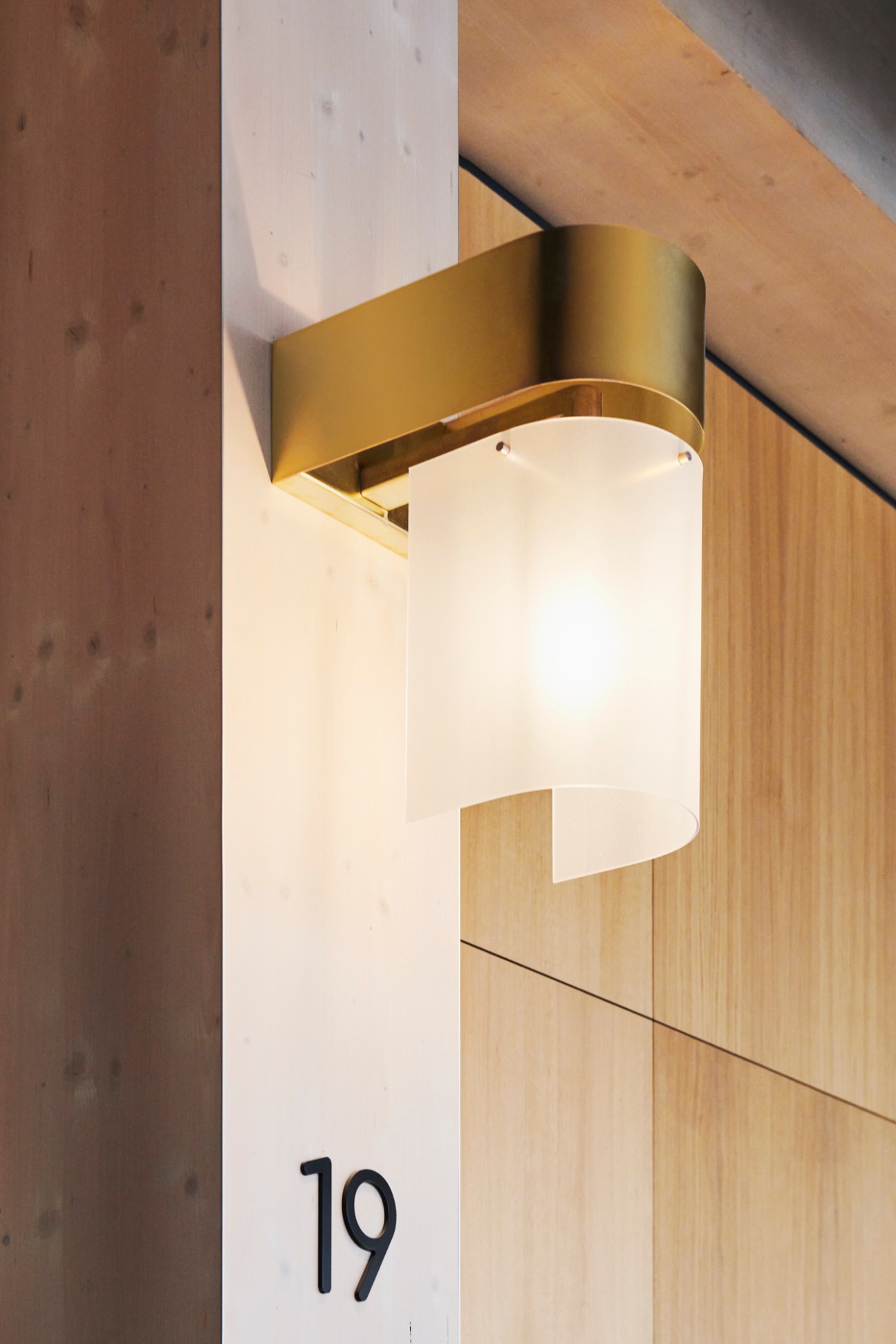 Modern cylindrical brass wall sconce with white lampshade mounted on light wood wall next to number 19