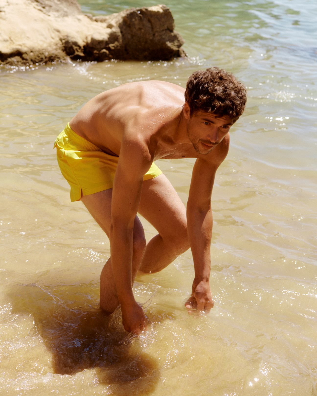 Man in yellow swim shorts crouching by water's edge at beach