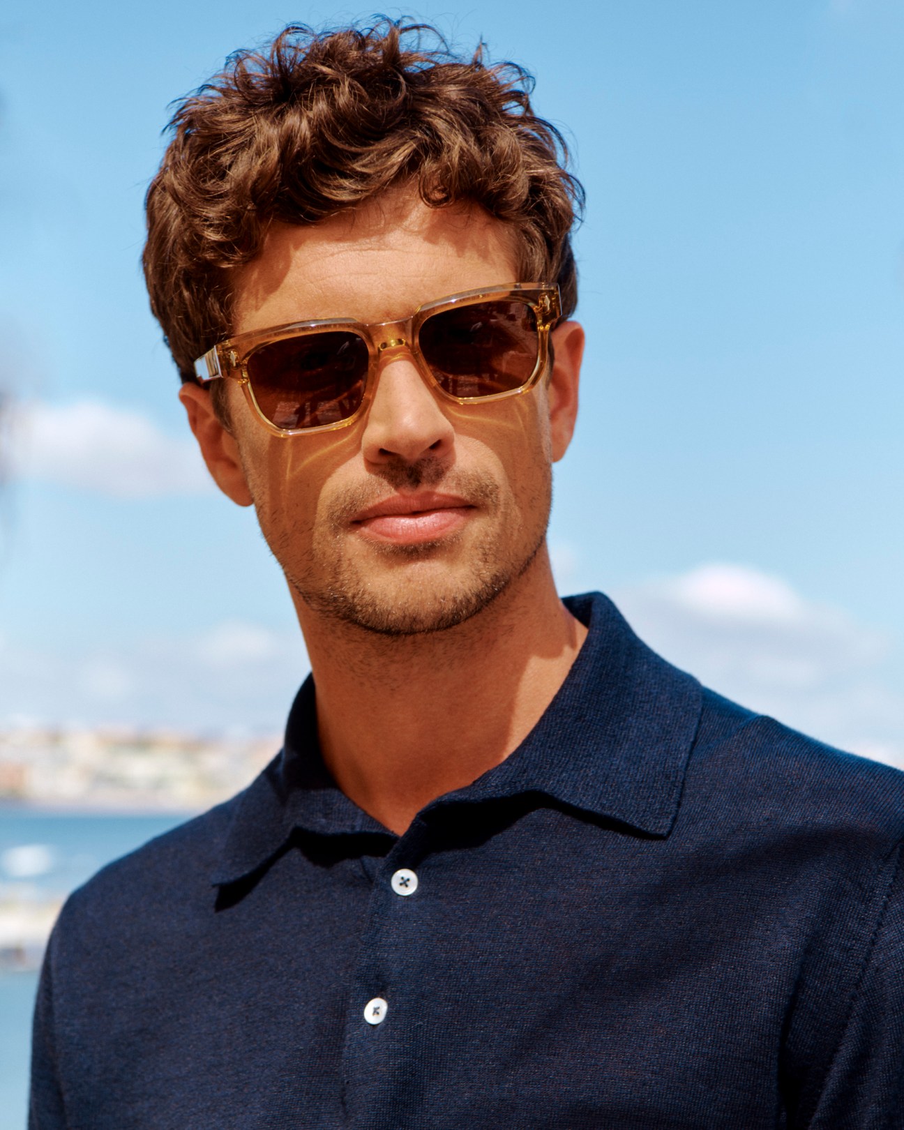 Man in navy polo shirt wearing translucent amber sunglasses against blue sky with coastline