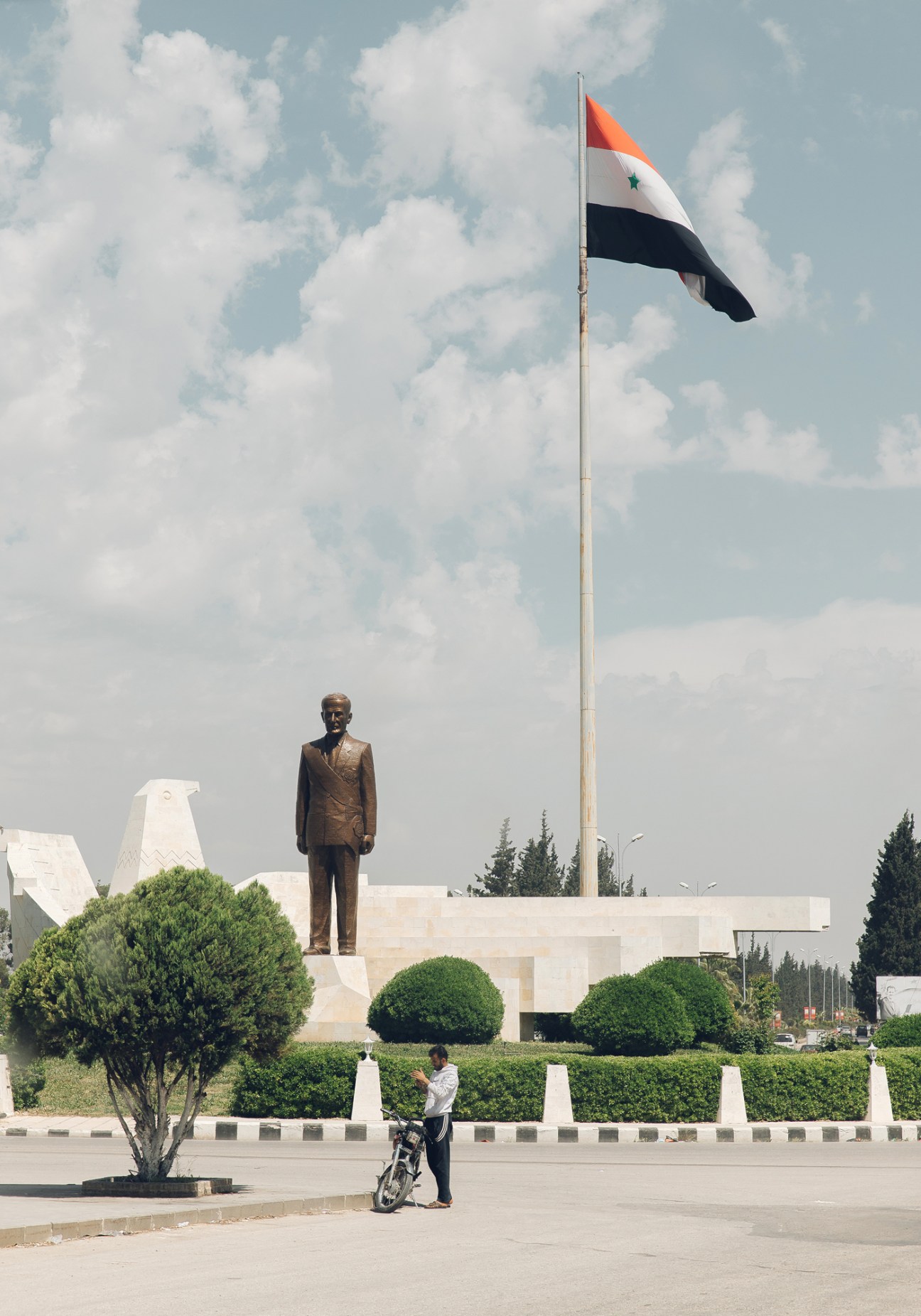 Statue of former Syrian president Hafez al-Assad on a white stone pedestal with Syrian flag on tall flagpole