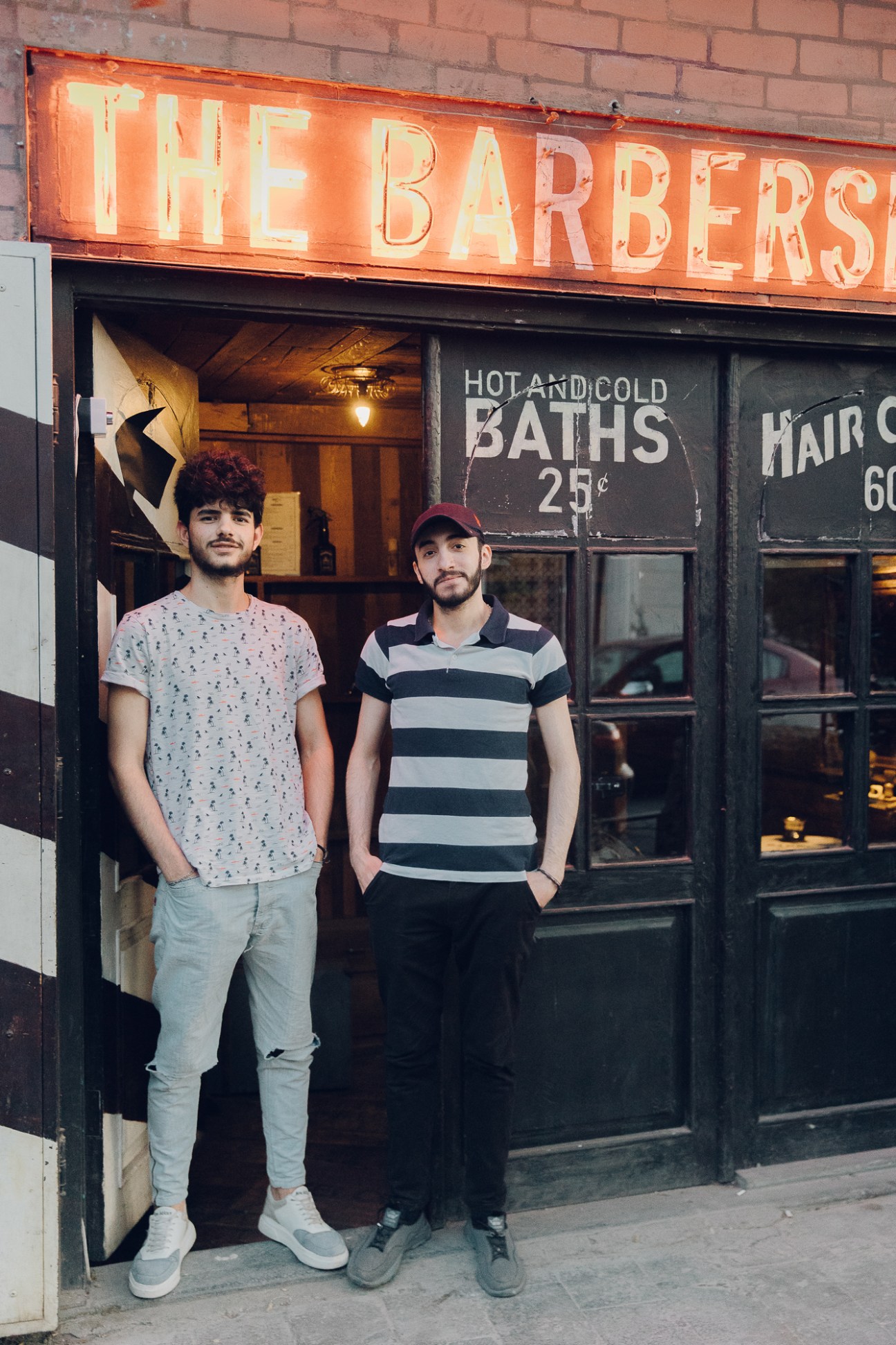 Two young men standing outside a vintage barbershop storefront with neon 'THE BARBERS' sign and painted window advertisements