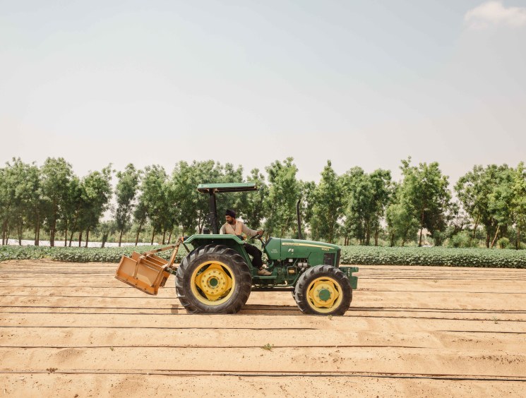 A farmer operates a green tractor with yellow wheels across sandy farmland at Emirates Bio Farm, with trees lining the background.