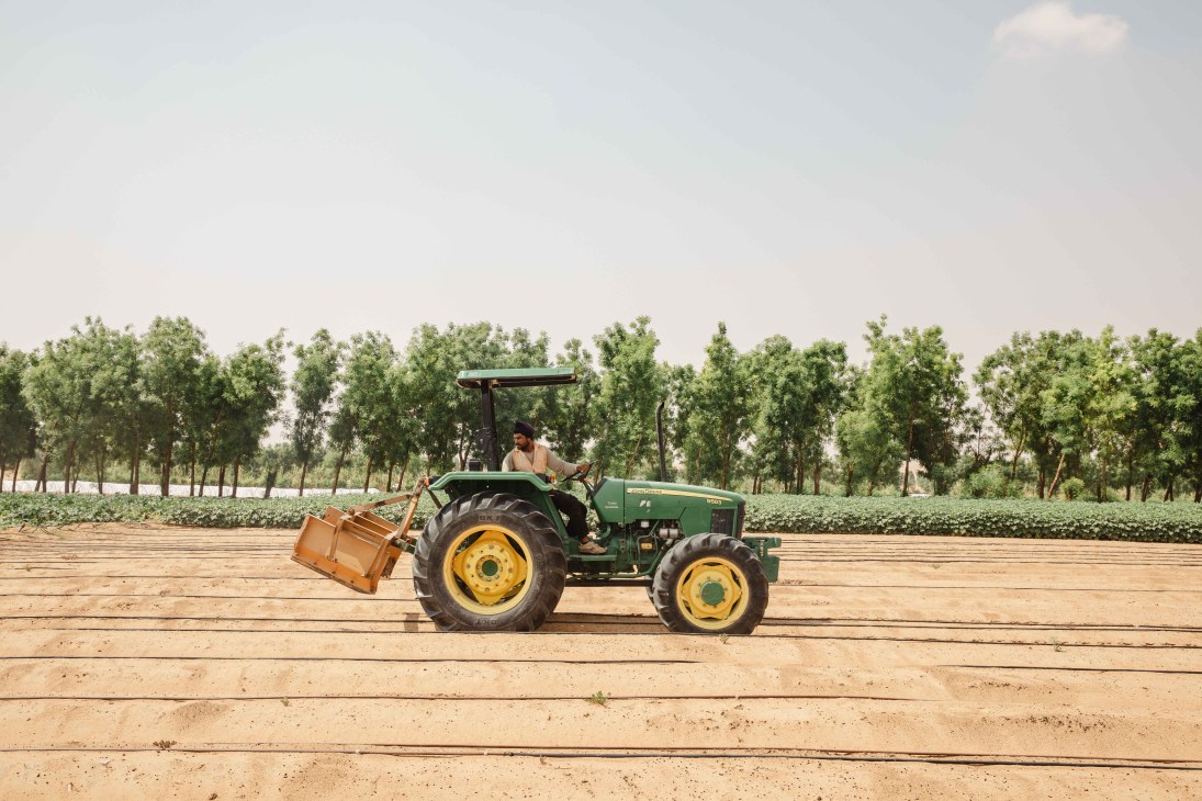 A farmer operates a green tractor with yellow wheels across sandy farmland at Emirates Bio Farm, with trees lining the background.