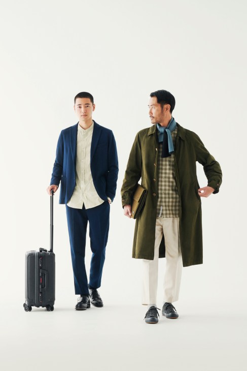 Two men in smart casual clothing posing against a white background, one in navy suit with suitcase, the other in olive coat and cream trousers.