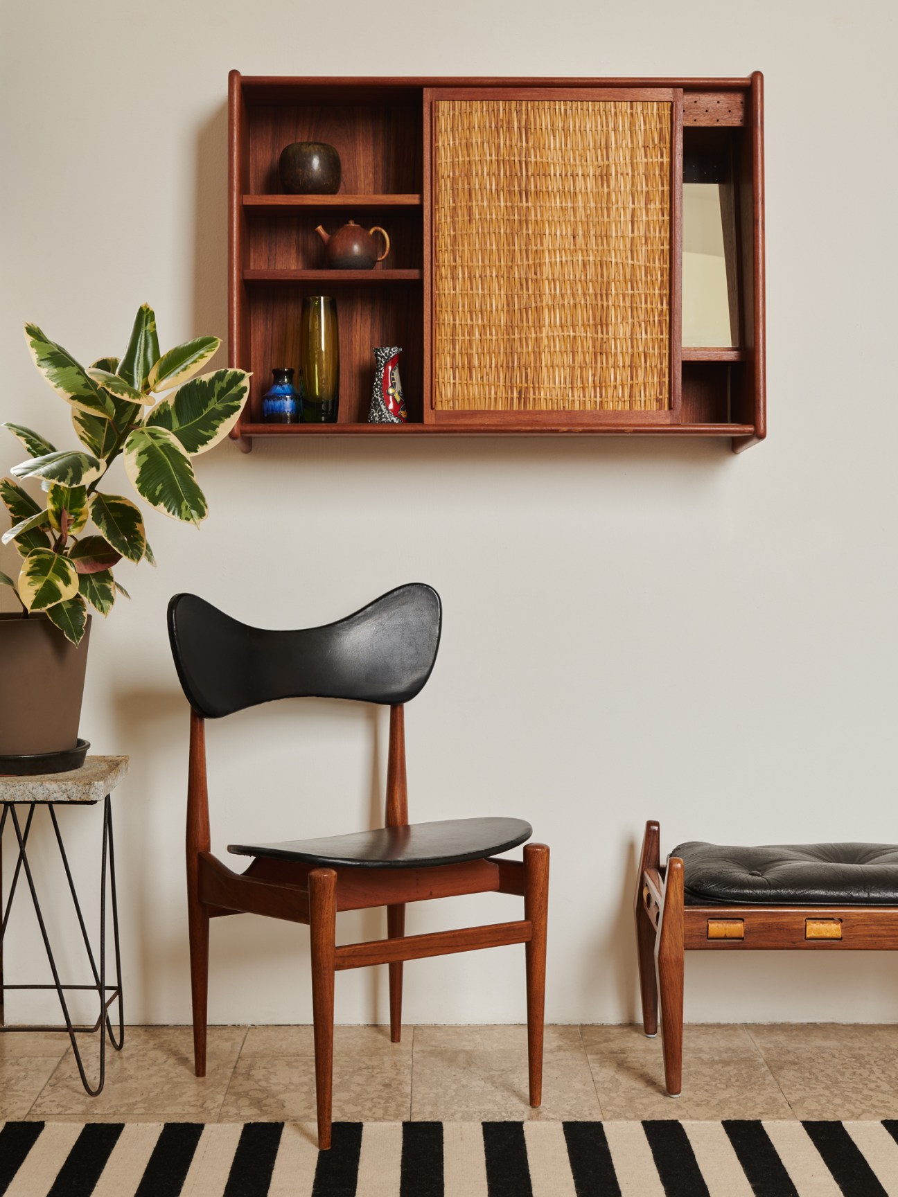 Mid-century modern wooden wall-mounted cabinet with shelving, decorative objects and woven panel door above a black leather chair.