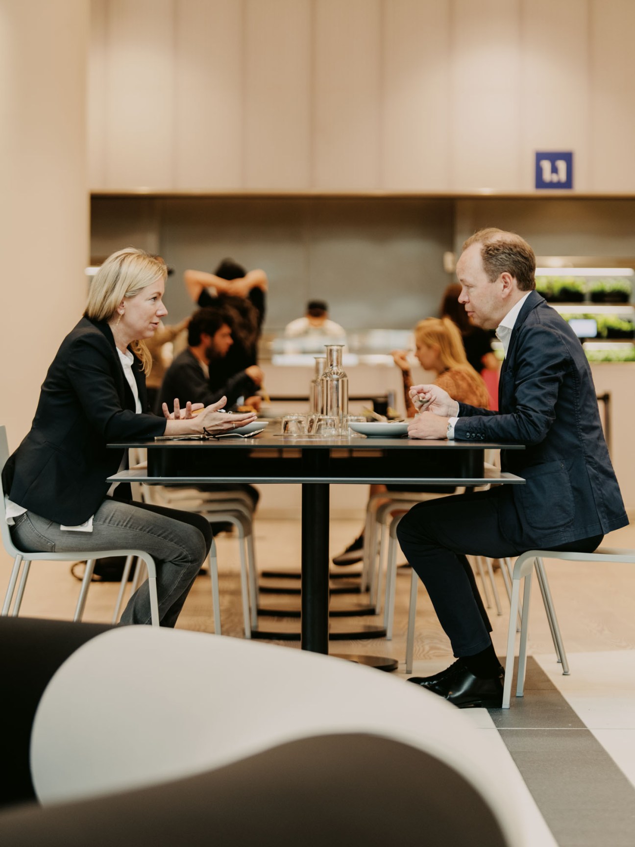 Stephanie Caspar and Julian Deutz in business attire having a conversation at a table in a modern office cafeteria