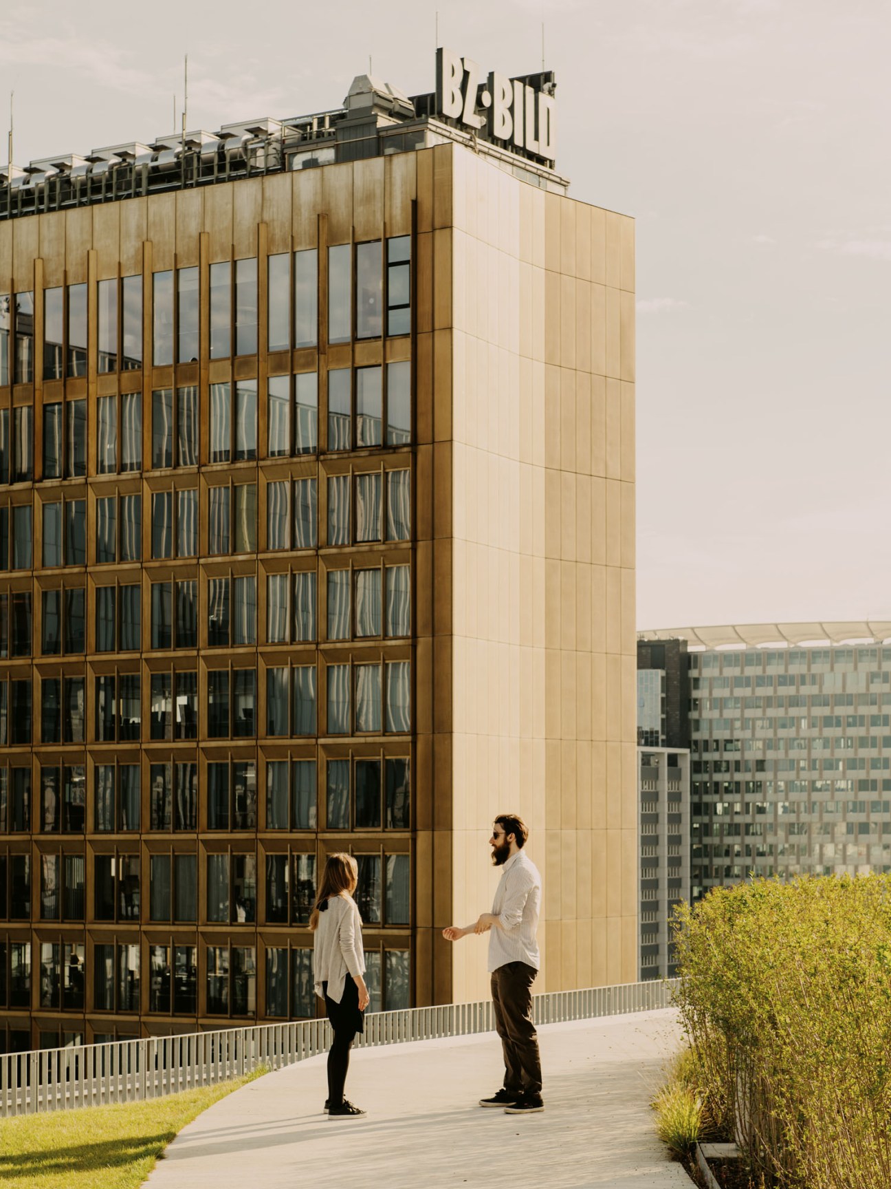 Two people having conversation on rooftop terrace with golden Axel Springer skyscraper visible in background