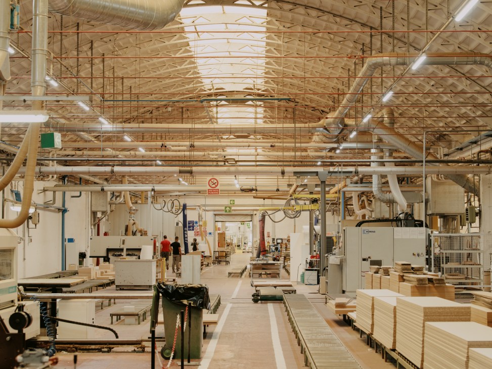 Industrial workshop with exposed ceiling beams, machinery, stacks of wood materials, and workers in a furniture factory