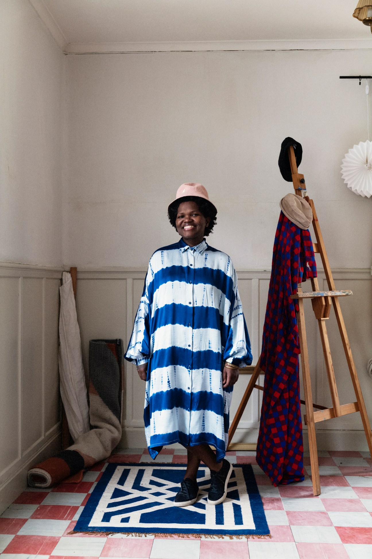 Nkuli Mlangeni-Berg standing in studio with blue and white striped garment and pink hat, textile samples visible