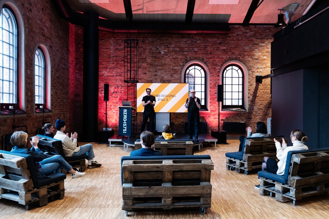Industry experts give a lecture to students seated on pallet furniture in Berghs School's industrial-style assembly hall with red brick walls and arched windows.