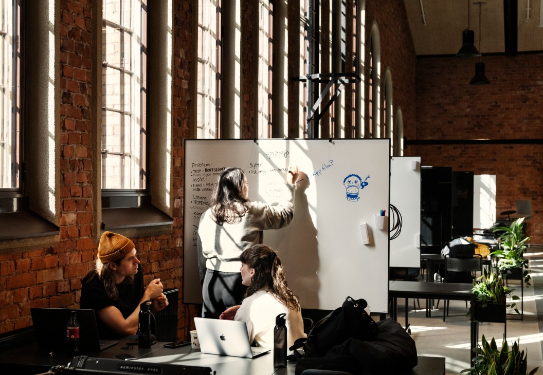Students collaborate at a whiteboard in Berghs School's industrial assembly hall with exposed red brick and large windows.