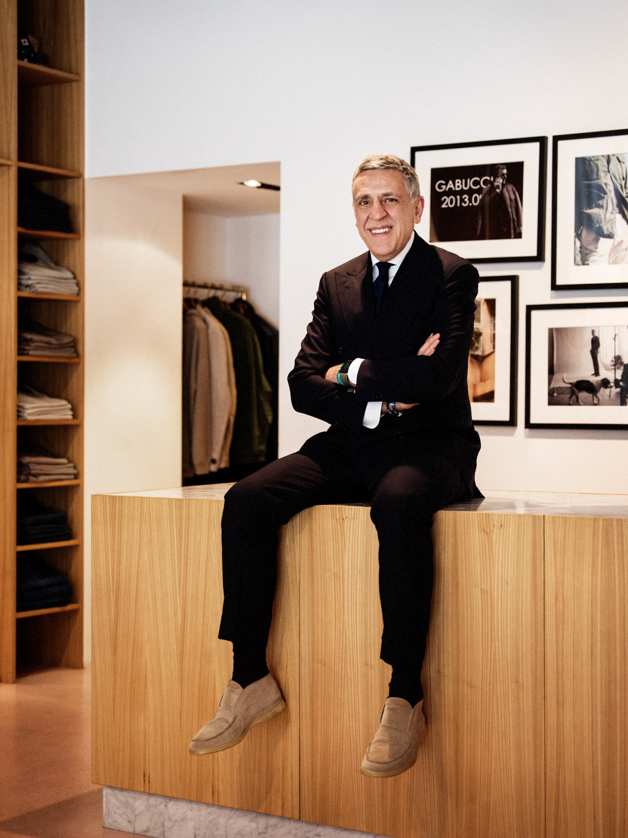 Bob Tavakoli sitting on wooden counter in his Gabucci menswear store with suits and shelving visible