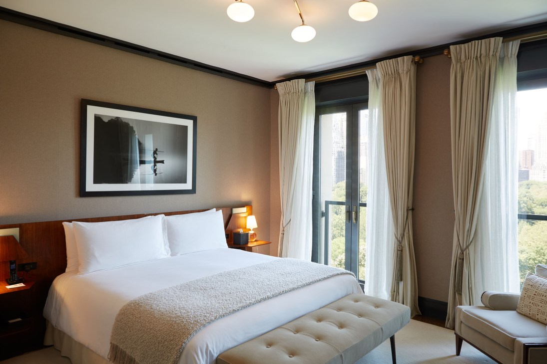 Elegant hotel bedroom with white bedding, neutral tones, large windows with curtains, and city view