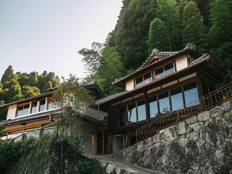 Traditional Japanese inn with curved tile roof nestled among dense green forest trees.