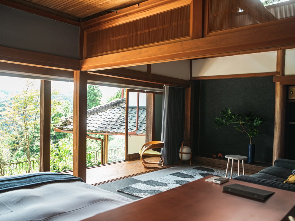 Modern Japanese inn room with wooden beams, sliding doors opening to forest views, bed, and minimalist furnishings.