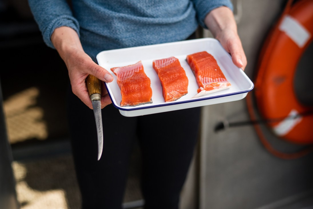 Person in blue sweater holding white enamel dish with three raw salmon fillets and a knife
