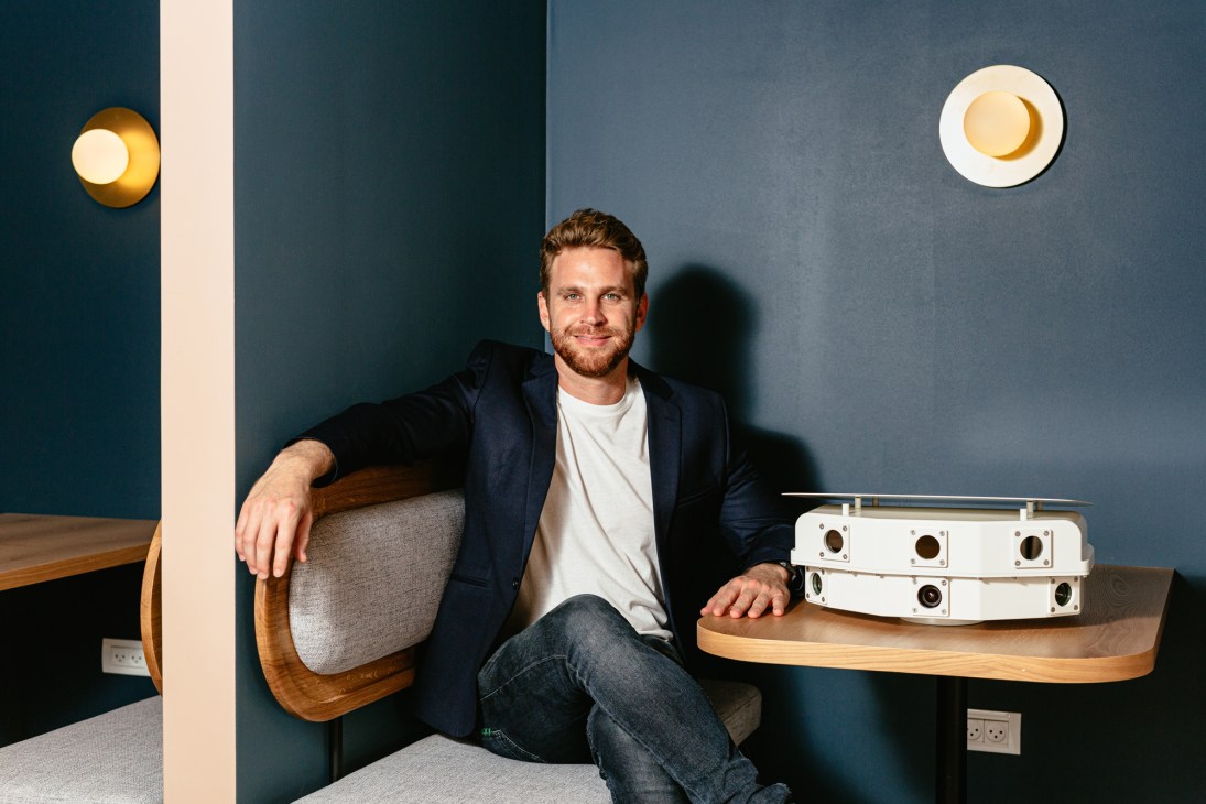 Man in dark blazer and white t-shirt sitting at wooden table with white audio equipment, against blue wall with gold wall sconces.