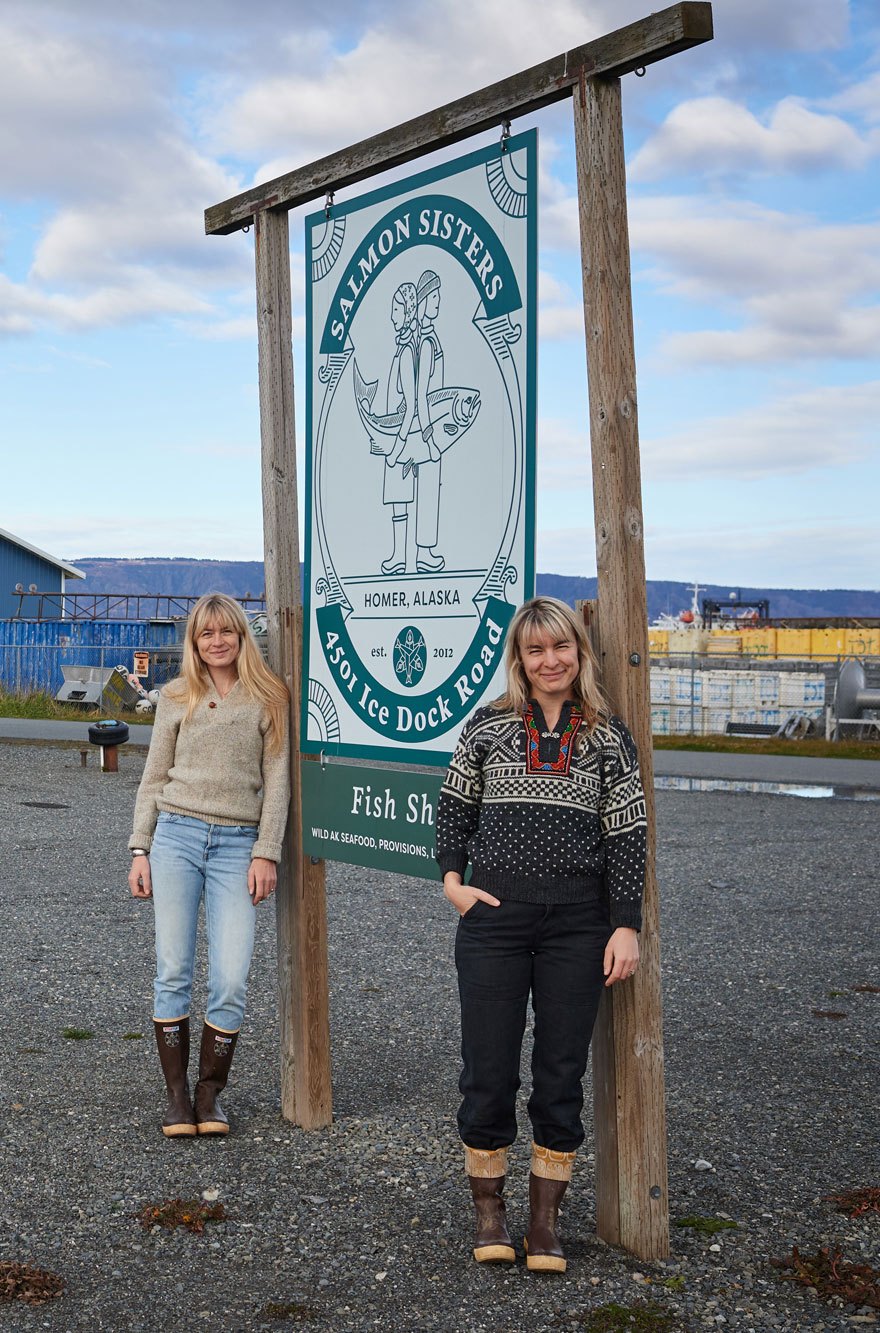 Two women in winter clothing standing beside a wooden sign for Salmon Sisters in Homer, Alaska