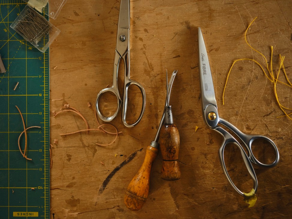 Leatherworking tools including scissors, awls, and cutting implements arranged on a wooden work surface