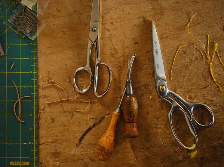 Leatherworking tools including scissors, awls, and cutting implements arranged on a wooden work surface