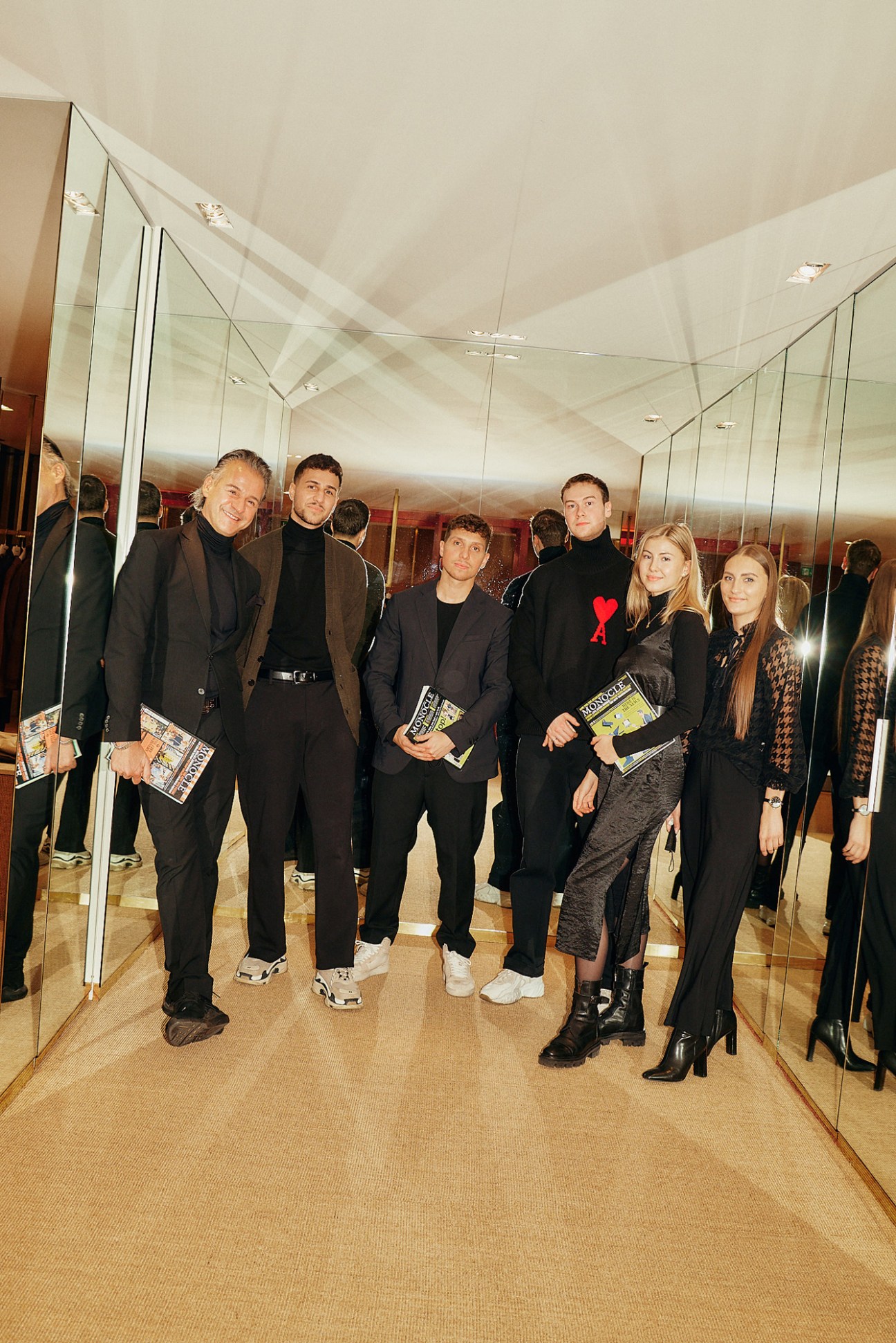 Group of five people in dark clothing posing together in a mirrored corridor holding magazines