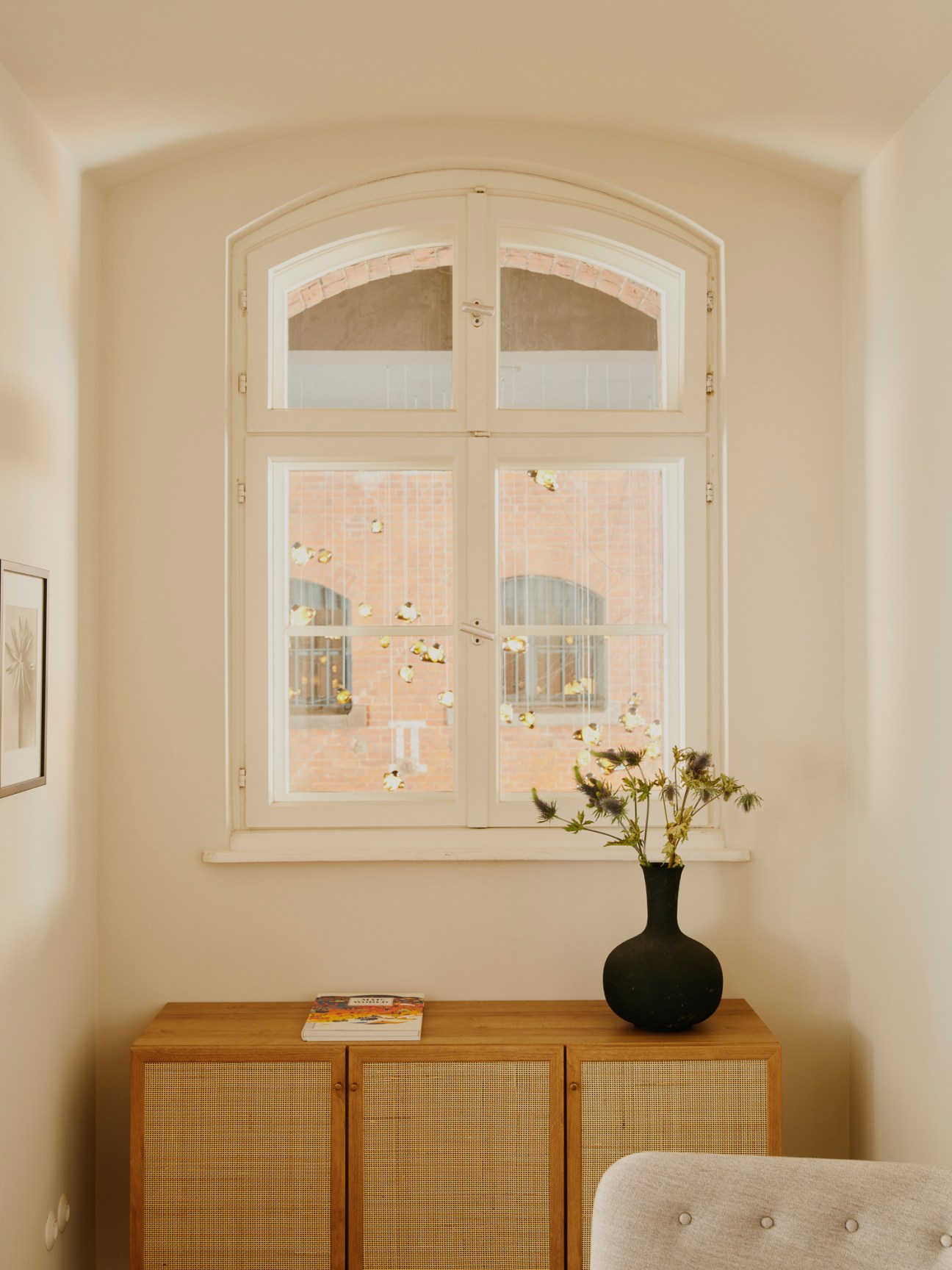 Hotel room interior with arched window, white walls, wooden sideboard and black vase with flowers