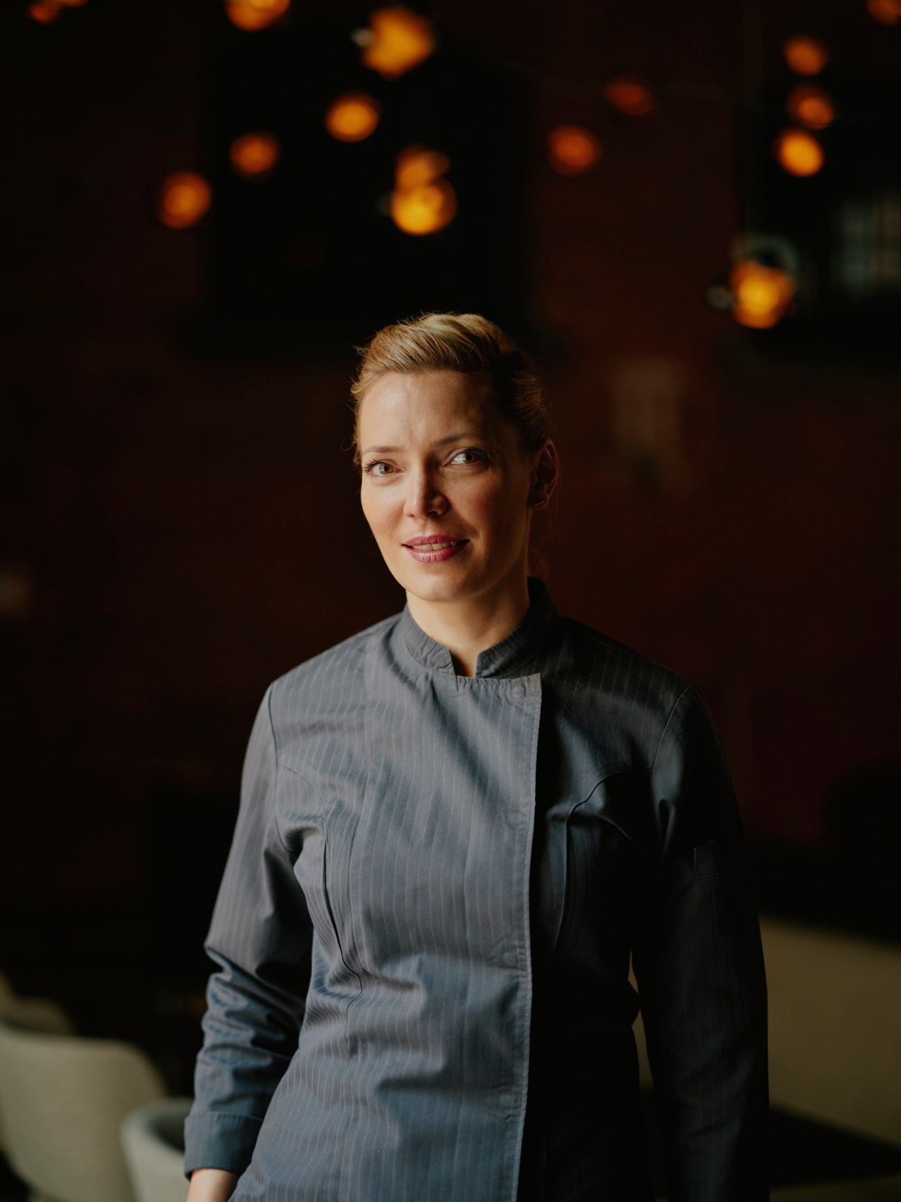 Chef Sophia Rudolph in chef's coat standing in restaurant with warm lighting in background