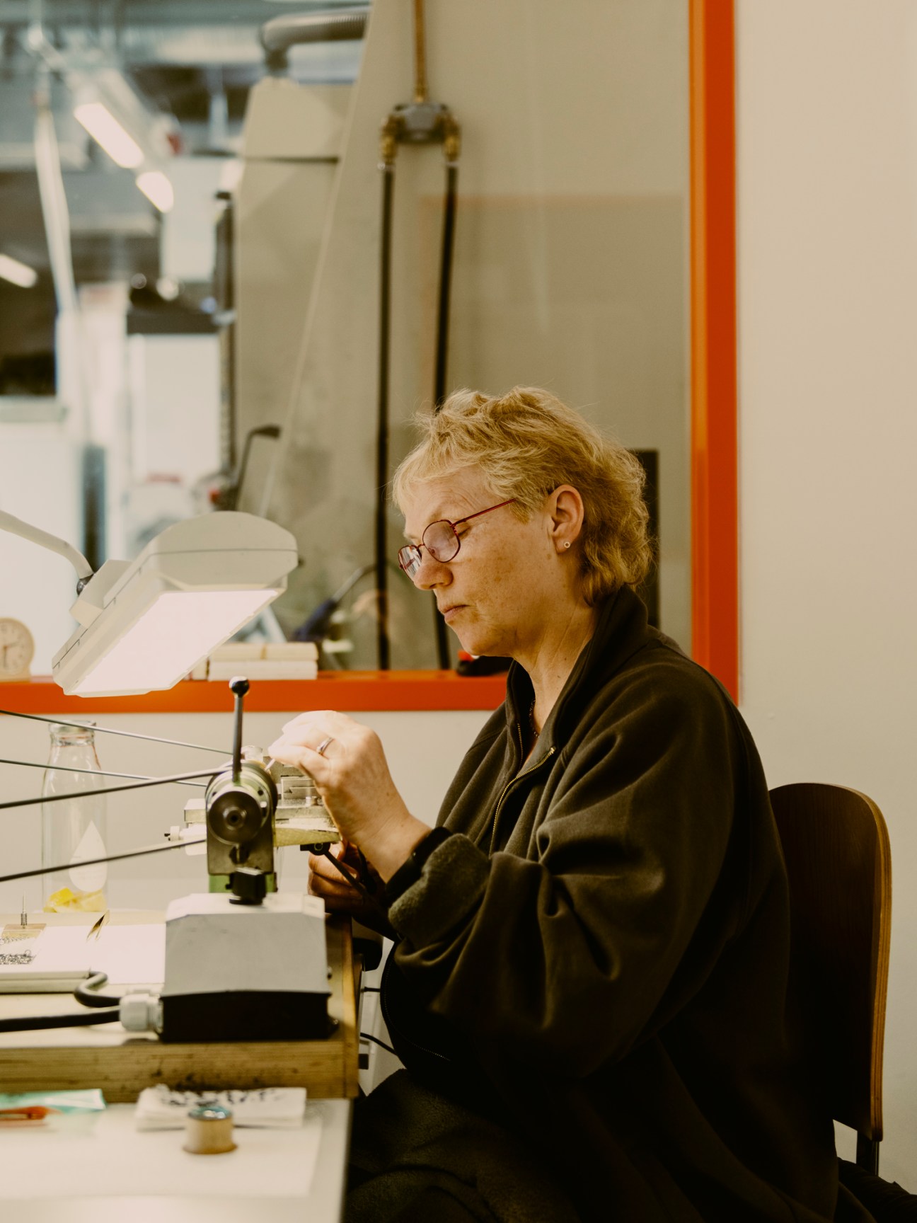 Watch craftsperson in glasses working with precision tools on intricate watch components at workbench