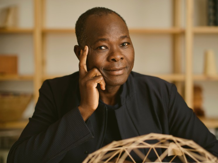 Diébédo Francis Kéré seated at his desk in his Berlin studio with a woven basket structure